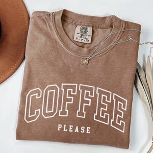 Peut inclure: T-shirt marron avec l'inscription "COFFEE PLEASE" en lettres blanches. Un collier argenté avec un petit pendentif repose sur le t-shirt. Un chapeau marron est visible dans le coin supérieur gauche.