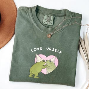 Pu&ograve; includere: T-shirt verde muschio con le parole "LOVE URSELF" sopra un cuore rosa contenente due rane dei cartoni animati con cappelli bianchi. Una collana d'oro con un ciondolo &egrave; sulla maglietta. Un cappello marrone &egrave; sullo sfondo.