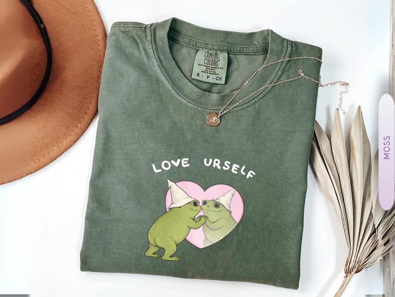 Pu&ograve; includere: T-shirt verde muschio con le parole "LOVE URSELF" sopra un cuore rosa contenente due rane dei cartoni animati con cappelli bianchi. Una collana d'oro con un ciondolo &egrave; sulla maglietta. Un cappello marrone &egrave; sullo sfondo.