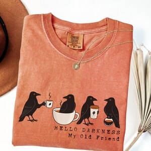 Peut inclure: T-shirt corail avec un motif de quatre corbeaux noirs, chacun tenant une tasse de café ou assis dans une tasse à café. Le texte "HELLO DARKNESS My Old Friend" est imprimé en dessous. Un collier doré et un chapeau marron sont également visibles.