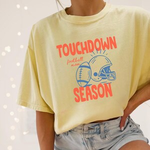 Puede incluir: Camiseta amarilla clara con las palabras "TOUCHDOWN SEASON" en coral, junto con un gr&aacute;fico de f&uacute;tbol americano y casco azul. La camiseta tambi&eacute;n tiene las palabras "football mom". La modelo lleva un pantal&oacute;n corto vaquero.