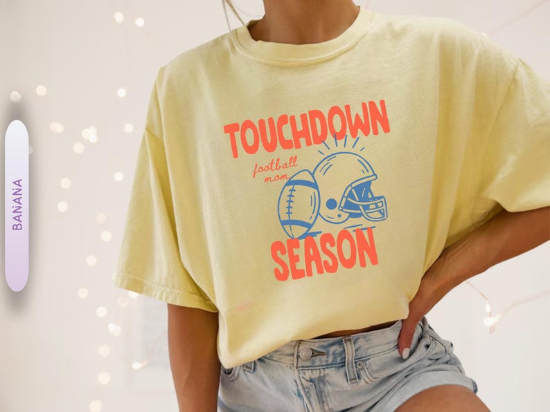 Puede incluir: Camiseta amarilla clara con las palabras "TOUCHDOWN SEASON" en coral, junto con un gr&aacute;fico de f&uacute;tbol americano y casco azul. La camiseta tambi&eacute;n tiene las palabras "football mom". La modelo lleva un pantal&oacute;n corto vaquero.