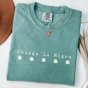 May include: A light green t-shirt with the text "Chinga la Migra" and four daisy graphics. A silver necklace with a pendant rests on the shirt. The shirt is labeled "Comfort Colors" and is a size small.
