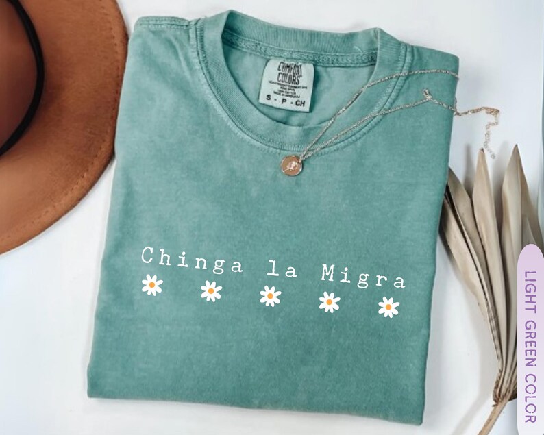 May include: A light green t-shirt with the text "Chinga la Migra" and four daisy graphics. A silver necklace with a pendant rests on the shirt. The shirt is labeled "Comfort Colors" and is a size small.