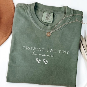 May include: An olive green t-shirt with the text "GROWING TWO TINY humans" and baby footprints. A gold necklace with a pendant rests on the shirt. A brown hat and dried plants are in the background. The shirt is labeled "Moss Color".