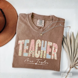 Peut inclure: Un t-shirt marron à manches courtes avec le mot TEACHER imprimé dans un motif patchwork floral et vichy. Sous le mot, le texte Mrs. Taylor est imprimé en écriture blanche avec un petit cœur. Le t-shirt porte une étiquette de marque Comfort Colors.