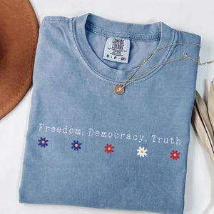 May include: A blue jean colored t-shirt with the words "Freedom, Democracy, Truth" printed on the front, along with flower graphics. A gold necklace and a brown hat are also visible.