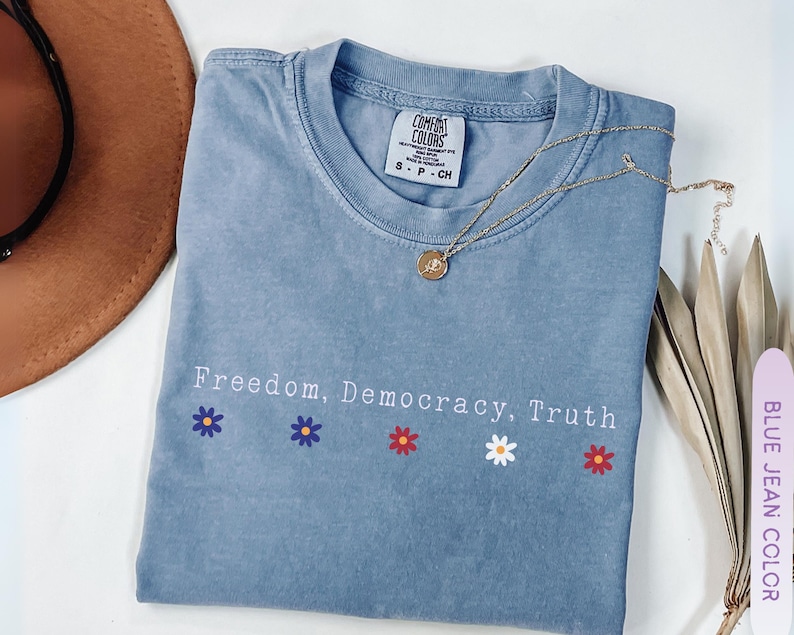 May include: A blue jean colored t-shirt with the words "Freedom, Democracy, Truth" printed on the front, along with flower graphics. A gold necklace and a brown hat are also visible.