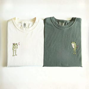May include: Two crew neck t-shirts in ivory and moss green. The ivory shirt features a small embroidered frog with a pink bow and three red hearts. The moss green shirt features a small embroidered frog holding a pink flower. Both have labels.