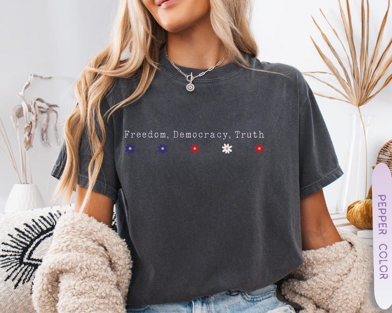 May include: Dark gray t-shirt with the words "Freedom, Democracy, Truth" printed across the chest. The shirt features small embroidered flowers in blue, blue, red, white, and red. The shirt is made of a soft, comfortable material.