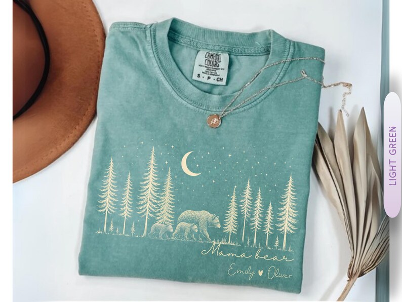 May include: A light green short-sleeve t-shirt featuring a cream-colored graphic of a forest with pine trees, a crescent moon, stars, and a bear with two cubs. The text reads Mama bear followed by the names Emily and Oliver with a heart symbol.