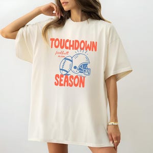 Puede incluir: Camiseta de color marfil con las palabras "TOUCHDOWN SEASON" en rojo, junto con un bal&oacute;n de f&uacute;tbol y un gr&aacute;fico de casco en azul. Las palabras "football mom" est&aacute;n encima del gr&aacute;fico. La camiseta es de corte holgado.