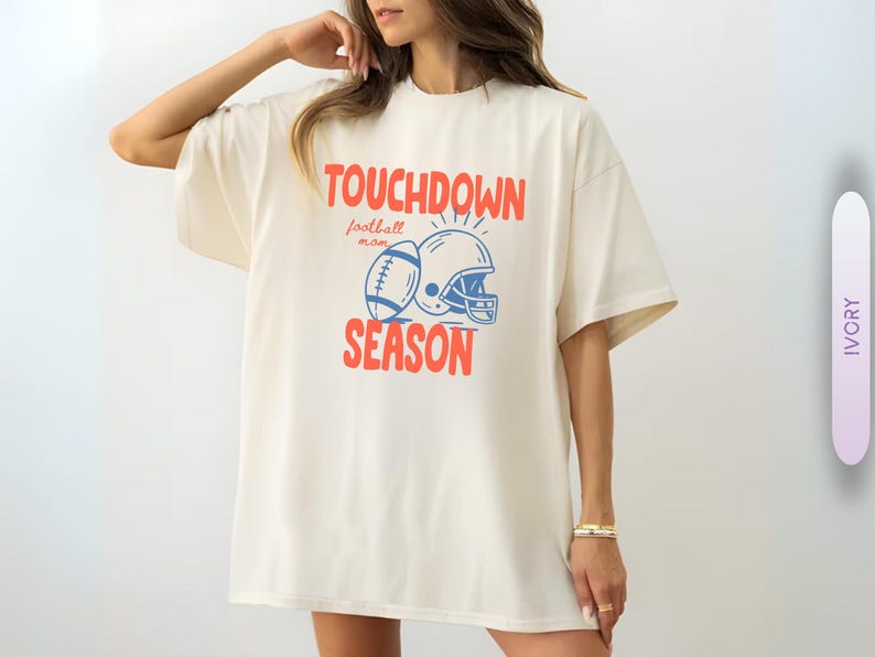 Puede incluir: Camiseta de color marfil con las palabras "TOUCHDOWN SEASON" en rojo, junto con un bal&oacute;n de f&uacute;tbol y un gr&aacute;fico de casco en azul. Las palabras "football mom" est&aacute;n encima del gr&aacute;fico. La camiseta es de corte holgado.