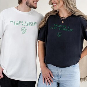 K&ouml;nnte beinhalten: Zwei T-Shirts, eines wei&szlig; und eines schwarz, mit gr&uuml;nem Text und Grafiken. Das wei&szlig;e Shirt sagt "DAD MODE LOADING NEED GUINNESS" mit einer Bierkrug-Grafik. Das schwarze Shirt sagt "GROWING A TINY SHAMROCK" mit einem Kleeblatt.