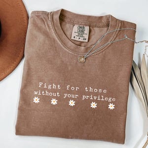 May include: Espresso-colored t-shirt with the text "Fight for those without your privilege" and daisy graphics. A silver necklace with a pendant rests on the shirt. A brown hat and decorative leaf are also visible.