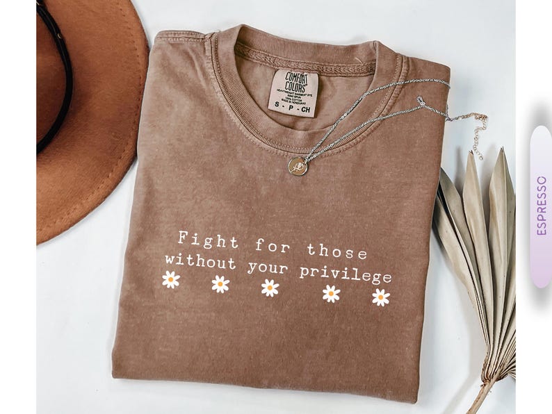 May include: Espresso-colored t-shirt with the text "Fight for those without your privilege" and daisy graphics. A silver necklace with a pendant rests on the shirt. A brown hat and decorative leaf are also visible.