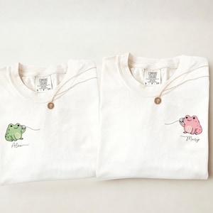 Peut inclure: Deux t-shirts ivoire à col rond. Chaque chemise présente un dessin de grenouille de dessin animé, une verte et une rose, tenant un téléphone. Le nom "Alex" est sous la grenouille verte et "Mary" sous la grenouille rose. Chaque chemise a un collier en or.