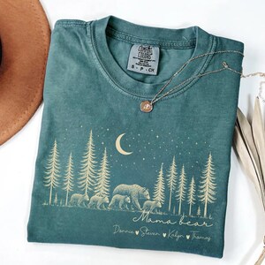 May include: A blue spruce colored t-shirt featuring a gold graphic of a bear family walking through a pine forest under a crescent moon and stars. The text reads Mama bear followed by the names Donnie, Steven, Kalyn, and Thomas with heart icons.