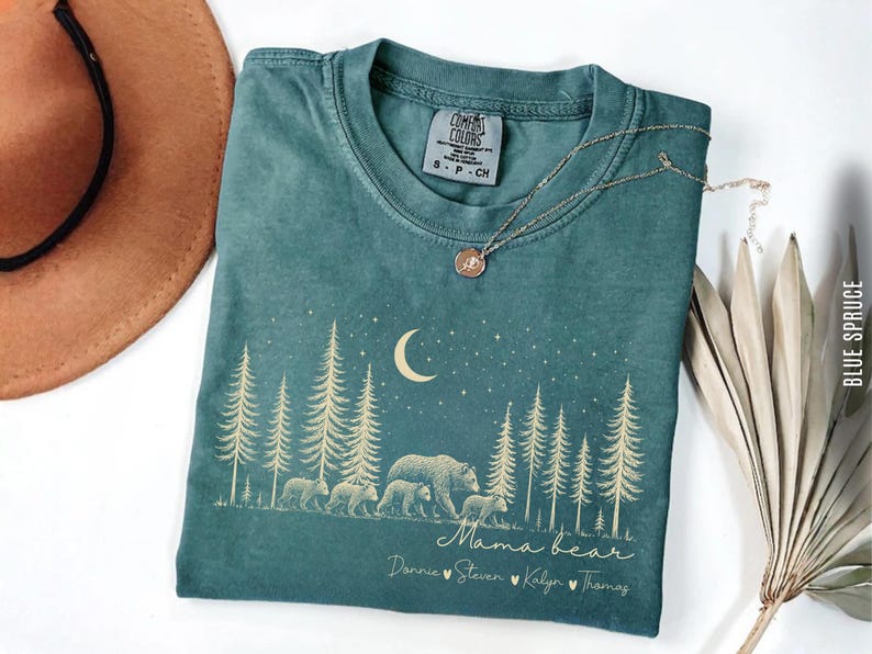 May include: A blue spruce colored t-shirt featuring a gold graphic of a bear family walking through a pine forest under a crescent moon and stars. The text reads Mama bear followed by the names Donnie, Steven, Kalyn, and Thomas with heart icons.