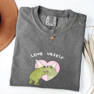 Pu&ograve; includere: T-shirt grigio scuro con le parole "LOVE URSELF" sopra una grafica di due rane verdi con cappelli bianchi all'interno di un cuore rosa. Sono visibili anche una collana d'oro e un cappello marrone.