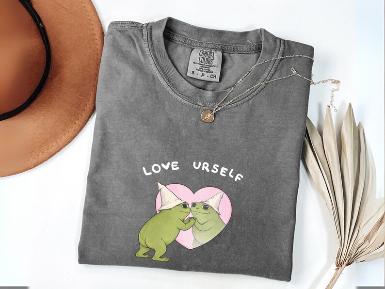 Pu&ograve; includere: T-shirt grigio scuro con le parole "LOVE URSELF" sopra una grafica di due rane verdi con cappelli bianchi all'interno di un cuore rosa. Sono visibili anche una collana d'oro e un cappello marrone.