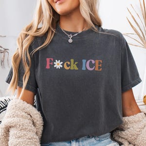 May include: Dark gray t-shirt with the text "F*ck ICE" in colorful, retro-style lettering. The word "F*ck" is in pink, the asterisk is a white daisy, and the word "ICE" is in blue and purple. The shirt is a casual, unisex style.