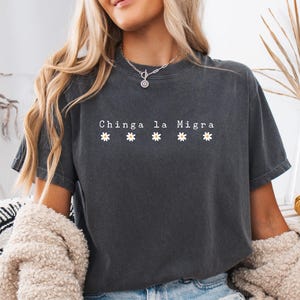 May include: Dark gray t-shirt with the text "Chinga la Migra" and five small daisy graphics. The shirt is a short-sleeved, crew neck style. The model is wearing a cream-colored cardigan.
