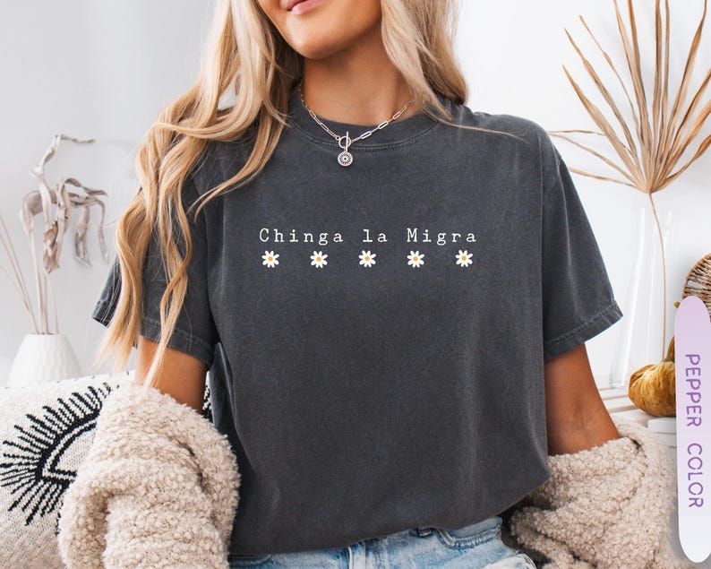 May include: Dark gray t-shirt with the text "Chinga la Migra" and five small daisy graphics. The shirt is a short-sleeved, crew neck style. The model is wearing a cream-colored cardigan.