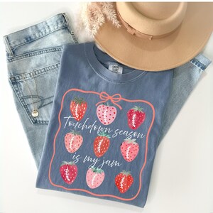 Peut inclure: Un t-shirt bleu avec un motif de fraises et le texte "Touchdown season is my jam". Le t-shirt est associé à un jean bleu clair et un chapeau beige. Un collier en or est également visible.