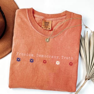 May include: Terracotta-colored t-shirt with the words "Freedom, Democracy, Truth" and flower graphics. A gold necklace with a pendant rests on the shirt. A brown hat and dried plant are in the background.