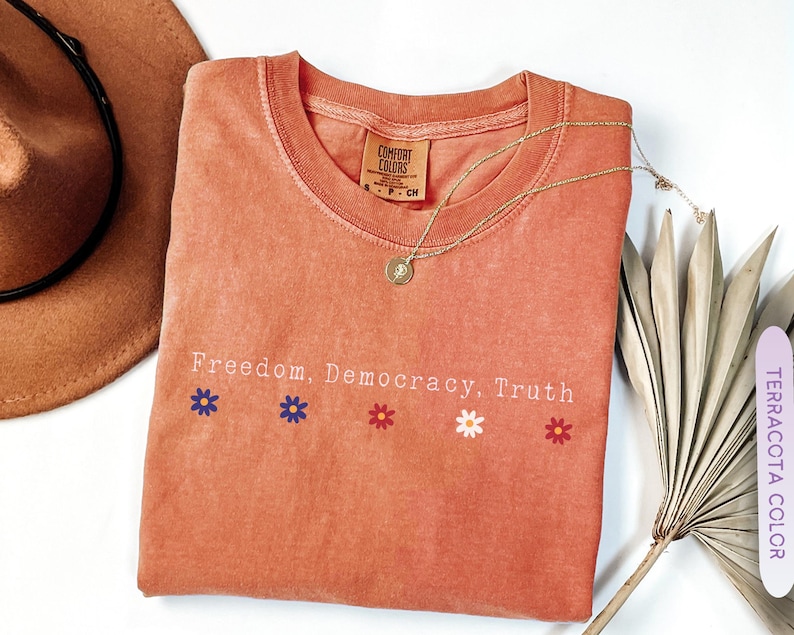 May include: Terracotta-colored t-shirt with the words "Freedom, Democracy, Truth" and flower graphics. A gold necklace with a pendant rests on the shirt. A brown hat and dried plant are in the background.