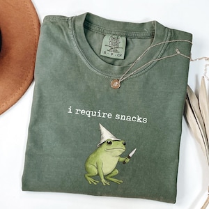 May include: A moss green t-shirt with the text "i require snacks" and a cartoon frog wearing a wizard hat and holding a knife. A brown hat and a gold necklace are also visible.