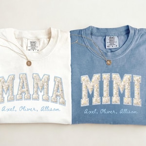 Peut inclure: Deux t-shirts pliés, un ivoire et un bleu, chacun avec un collier doré. Le t-shirt ivoire dit "MAMA" et le t-shirt bleu dit "MIMI", tous deux dans un motif floral. En dessous, les noms "Axel, Oliver, Allison".