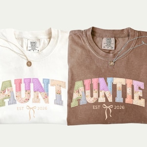 Peut inclure: Deux t-shirts pliés, un ivoire et un marron, chacun avec le mot "AUNTIE" en lettres patchwork colorées. En dessous, "EST 2026" et un motif de nœud. Chaque chemise a un collier.