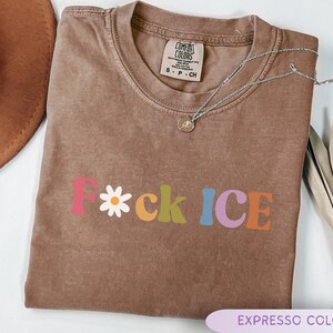 May include: A brown, short-sleeved t-shirt with the text "F*ck ICE" in colorful, retro-style lettering. The letter "F" is pink, the asterisk is a white daisy, and the other letters are in various colors. A silver necklace with a small pendant rests on the shirt.