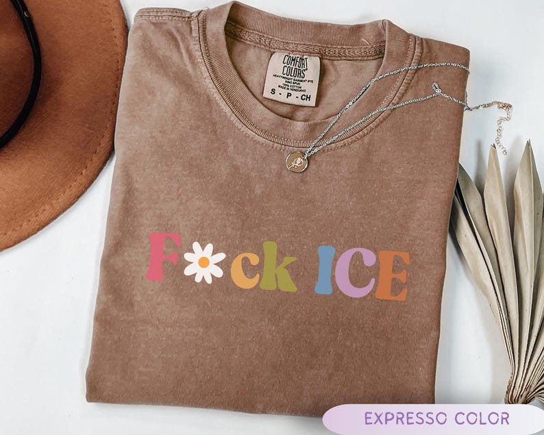 May include: A brown, short-sleeved t-shirt with the text "F*ck ICE" in colorful, retro-style lettering. The letter "F" is pink, the asterisk is a white daisy, and the other letters are in various colors. A silver necklace with a small pendant rests on the shirt.