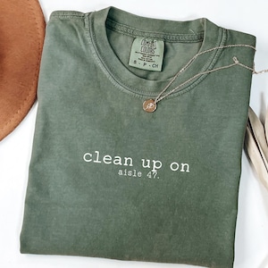 May include: An olive green t-shirt with the text "clean up on aisle 47" in white. A gold necklace with a pendant rests on the shirt. A brown hat and dried plants are in the background. A color swatch labeled "moss color" is also visible.