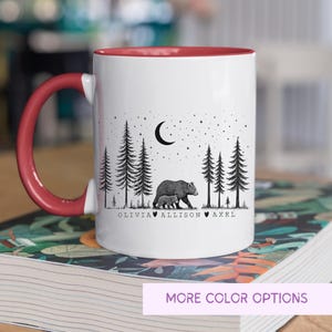 May include: White ceramic mug with a red interior and handle. The mug features a black and white illustration of a bear and cub in a forest under a crescent moon and stars. The names Olivia, Allison, and Axel are printed below the image.
