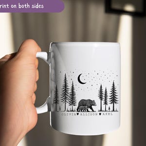 May include: White ceramic mug featuring a black and white illustration of a bear and cub in a forest under a crescent moon and stars. The mug has the text "Print on both sides" and names "Olivia, Allison, Axel" below the bear illustration.