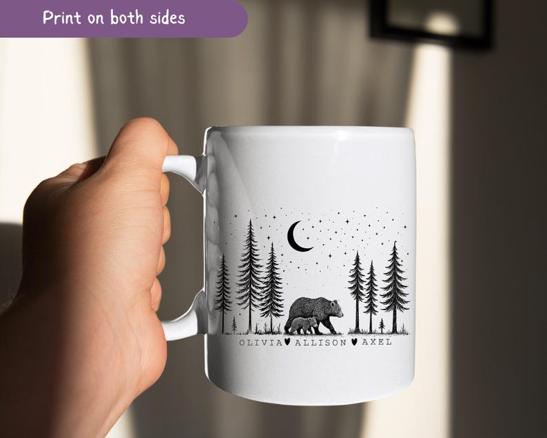 May include: White ceramic mug featuring a black and white illustration of a bear and cub in a forest under a crescent moon and stars. The mug has the text "Print on both sides" and names "Olivia, Allison, Axel" below the bear illustration.