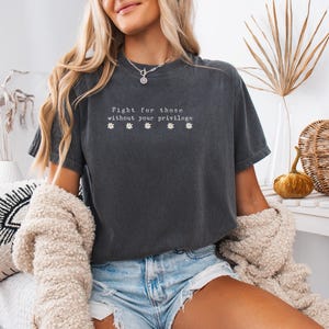 May include: Dark gray t-shirt with the text "Fight for those without your privilege" and four small flower graphics. The shirt is worn with denim shorts and a cream-colored sweater. The background includes decorative items.