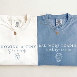 May include: Two folded t-shirts, one ivory and one blue, with text and graphics. The ivory shirt says "Growing a Tiny Shamrock" with a shamrock graphic. The blue shirt says "Dad Mode Loading. need Guinness" with a beer mug graphic.