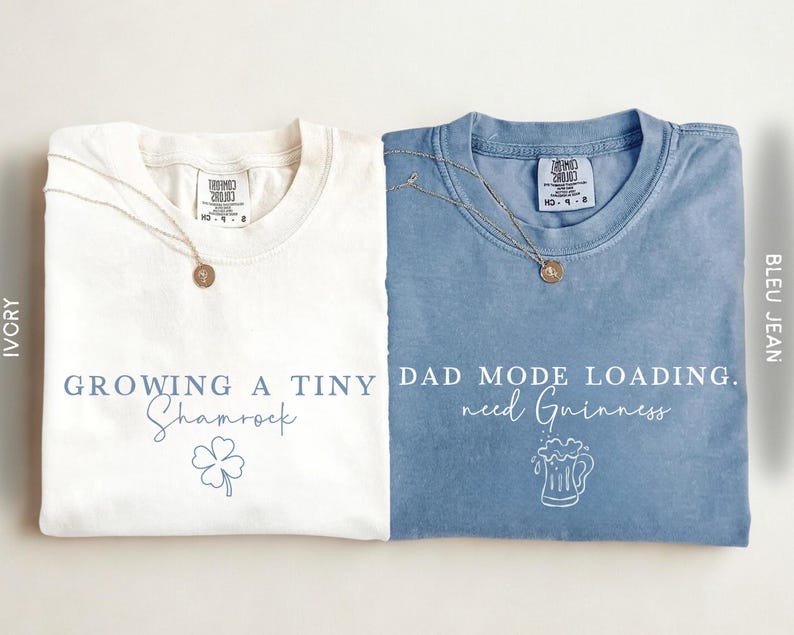 May include: Two folded t-shirts, one ivory and one blue, with text and graphics. The ivory shirt says "Growing a Tiny Shamrock" with a shamrock graphic. The blue shirt says "Dad Mode Loading. need Guinness" with a beer mug graphic.