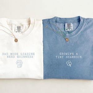 K&ouml;nnte beinhalten: Zwei gefaltete T-Shirts, eines in Elfenbein und eines in Blau, mit Texten und grafischen Designs. Das elfenbeinfarbene Shirt tr&auml;gt die Aufschrift "DAD MODE LOADING NEED GUINNESS" mit einer Bierkrug-Grafik. Das blaue Shirt sagt "GROWING A TINY SHAMROCK" mit einer Kleeblatt-Grafik. Beide haben Halsketten.