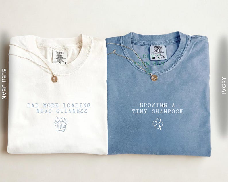 K&ouml;nnte beinhalten: Zwei gefaltete T-Shirts, eines in Elfenbein und eines in Blau, mit Texten und grafischen Designs. Das elfenbeinfarbene Shirt tr&auml;gt die Aufschrift "DAD MODE LOADING NEED GUINNESS" mit einer Bierkrug-Grafik. Das blaue Shirt sagt "GROWING A TINY SHAMROCK" mit einer Kleeblatt-Grafik. Beide haben Halsketten.
