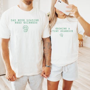 K&ouml;nnte beinhalten: Zwei wei&szlig;e T-Shirts mit gr&uuml;nem Text und Grafiken. Ein Shirt tr&auml;gt die Aufschrift "DAD MODE LOADING NEED GUINNESS" mit einer Bierkrug-Grafik. Das andere Shirt tr&auml;gt die Aufschrift "GROWING A TINY SHAMROCK" mit einer Kleeblatt-Grafik. Beide Shirts sind kurz&auml;rmelig.