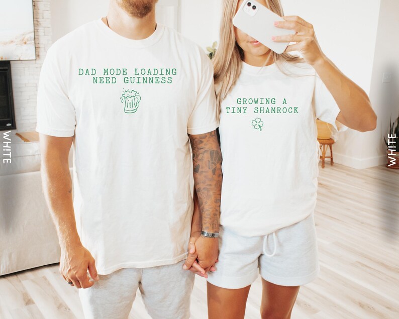 K&ouml;nnte beinhalten: Zwei wei&szlig;e T-Shirts mit gr&uuml;nem Text und Grafiken. Ein Shirt tr&auml;gt die Aufschrift "DAD MODE LOADING NEED GUINNESS" mit einer Bierkrug-Grafik. Das andere Shirt tr&auml;gt die Aufschrift "GROWING A TINY SHAMROCK" mit einer Kleeblatt-Grafik. Beide Shirts sind kurz&auml;rmelig.