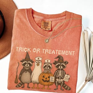 Peut inclure: T-shirt corail avec l'inscription "Trick or Treatement" et un motif de ratons laveurs et d'un oiseau en costumes d'Halloween. Le t-shirt a un col rond et une petite étiquette. Un collier doré et un chapeau marron sont également visibles.