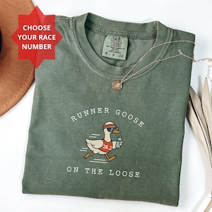 May include: A moss green crew neck t-shirt featuring a graphic of a goose wearing a red headband, sunglasses, and a race bib with the number 26.2, holding a water bottle. The text reads RUNNER GOOSE ON THE LOOSE and CHOOSE YOUR RACE NUMBER.