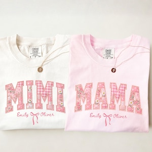 Peut inclure: Deux t-shirts, ivoire et rose, avec les mots "MIMI" et "MAMA" en lettres patchwork florales et vichy roses. Chaque chemise a un collier doré et les noms "Emily Oliver" en dessous.